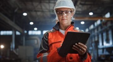 Eine Ingenieurin oder Technikerin in orangefarbener Sicherheitsjacke, weißem Schutzhelm und Schutzbrille arbeitet konzentriert auf einem Tab