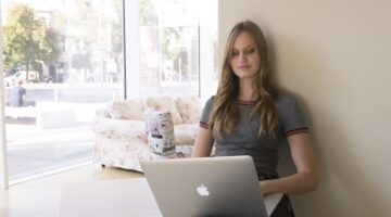 Eine junge Frau sitzt mit einem Apple MacBook auf dem Boden, an eine weiße Wand gelehnt, in einem he