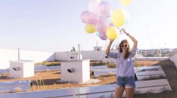 Eine junge Frau mit Sonnenbrille hält einen bunten Strauß aus Pastellballons über ihren Kopf und läc