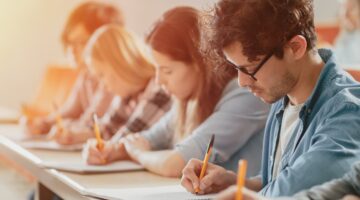 Mehrere Studierende sitzen in einer Reihe an Schreibtischen und schreiben konzentriert mit Bleistiften auf Papier, im Vordergrund ein junger