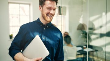 Ein lächelnder junger Mann mit rotem Bart und blauem Hemd hält einen Laptop unter dem Arm und geht d