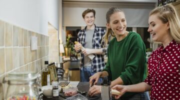 Drei junge Menschen kochen gemeinsam und lachen in einer gemütlichen WG-Küche, wobei zwei Frauen Käs