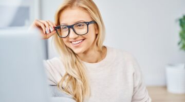 Eine junge blonde Frau mit schwarzer Brille lächelt selbstbewusst in die Kamera, während sie vor ein