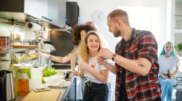 Vier junge Menschen kochen und lachen gemeinsam in einer modernen WG-Küche, während frisches Gemüse