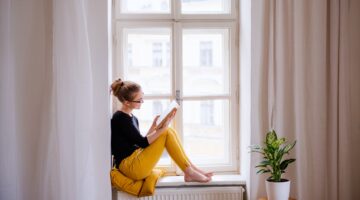 Eine junge Frau mit Brille und hochgesteckten Haaren sitzt barfuß auf einer Fensterbank und liest ei