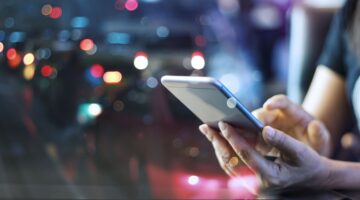 Eine Person hält ein Smartphone in beiden Händen und tippt darauf, während im unscharfen Hintergrund