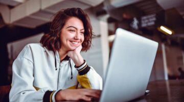 Eine lächelnde junge Frau mit kurzen braunen Haaren sitzt an einem Tisch und arbeitet entspannt an i