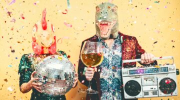 Zwei Personen in skurrilen Tiermasken (Dinosaurier und pinkes Fabelwesen) feiern ausgelassen mit Dis