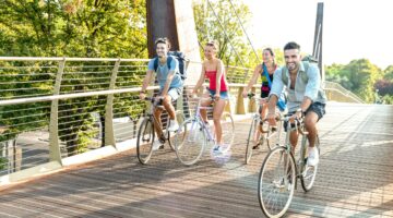 Vier fröhliche junge Menschen fahren gemeinsam auf Fahrrädern über eine moderne Holzbrücke mit Metal