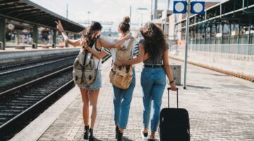 Drei junge Frauen mit Rucksäcken und einem Trolley laufen gemeinsam einen Bahnsteig entlang, eine he
