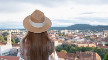 Eine junge Frau mit Strohhut und gemustertem Hemd blickt von einem Aussichtspunkt auf die Dächer von