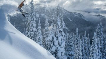Ein Skifahrer in orangefarbener Jacke springt über einen verschneiten Steilhang, umgeben von schneeb