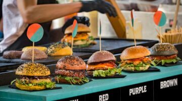 Vier bunte Gourmet-Burger mit unterschiedlich gefärbten Brötchen (gelb, rot, lila, braun) sind auf e