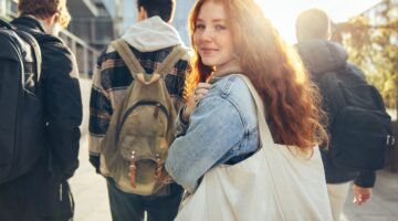 Eine junge rothaarige Studentin mit Jeansjacke und Tote Bag dreht sich lächelnd zur Kamera um, währe