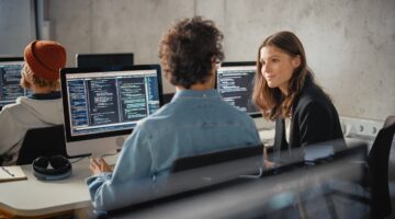 Zwei Studierende im Computerraum unterhalten sich während einer Programmierübung, auf den Monitoren