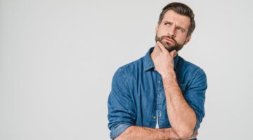Ein junger bärtiger Mann in einem blauen Jeanshemd hält nachdenklich die Hand ans Kinn und blickt sk