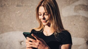 Eine junge Frau mit langen blonden Haaren lächelt entspannt auf ihr Smartphone, während sie vor eine