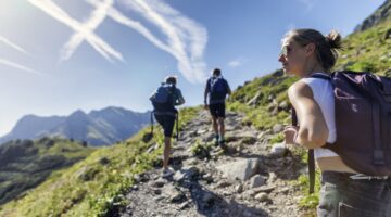 Drei junge Personen wandern auf einem felsigen Bergpfad aufwärts, im Hintergrund eine beeindruckende