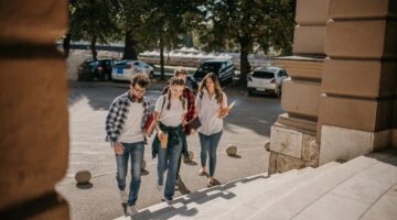 Vier junge Studierende gehen gemeinsam die Stufen eines Universitätsgebäudes hinauf, mit Büchern und