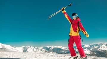 Eine lachende Skifahrerin in auffälliger rot-gelber Skiausrüstung reckt jubelnd ihre Skistöcke in di
