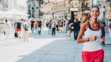 Eine junge, lächelnde Frau in roter Shorts und weißem Tank-Top joggt durch eine belebte Fußgängerzon