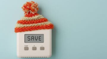 Ein weißer Thermostat mit dem Display-Text 'SAVE' trägt eine orangerot-weiß gestreifte Strickmütze m