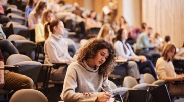 Eine junge Studentin mit lockigem Haar schreibt konzentriert Notizen in einem gut gefüllten Hörsaal