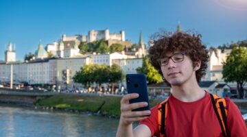 Ein junger Mann mit lockigem Haar und Brille schaut konzentriert auf sein Smartphone, im Hintergrund
