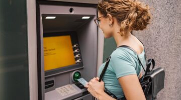 Eine junge Frau mit lockigem Haar, Brille und schwarzem Rucksack hebt am Geldautomaten Bargeld ab un