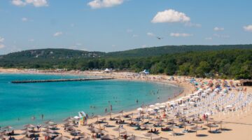 Belebter Sandstrand mit zahlreichen Sonnenschirmen und Liegestühlen an einer türkisblauen Bucht, umg