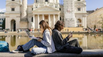 Zwei Studierende sitzen Rücken an Rücken auf einer Steinmauer vor der Wiener Karlskirche – eine jung