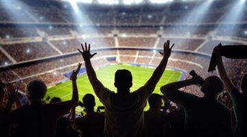 Jubelnde Fußballfans im Stadion als Silhouetten vor einem hell erleuchteten grünen Rasen, einer reiß