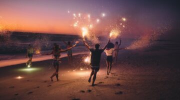 Eine Gruppe junger Menschen läuft ausgelassen am Strand entlang und schwenkt Wunderkerzen, während e