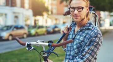 Ein junger blonder Mann mit Brille und Karohemd trägt sein Fahrrad auf der Schulter und lächelt ents