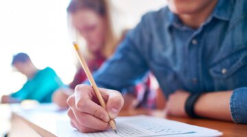 Ein Student füllt mit einem Bleistift einen Multiple-Choice-Fragebogen aus, im Hintergrund sind weit