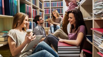 Fünf junge Studierende sitzen und stehen entspannt zwischen hohen Bücherregalen in einer Bibliothek,