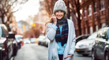Eine lächelnde junge Frau in winterlichem Outfit – grauer Strickmütze, buntem Schal und großem Cardi