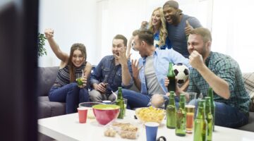 Eine fröhliche Gruppe von sechs jungen Menschen schaut gemeinsam ein Fußballspiel im Wohnzimmer und