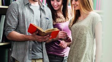 Drei junge Studierende – ein Mann mit Brille und zwei Frauen – stehen lächelnd in einer Bibliothek u