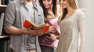 Drei lächelnde Studierende – ein junger Mann mit Brille und zwei junge Frauen – stehen gemeinsam in