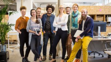 Eine diverse Gruppe von acht jungen Erwachsenen posiert lächelnd in einem modernen, hellen Startup-B
