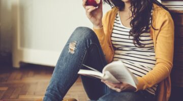 Eine junge Frau mit Brille sitzt entspannt auf einem gemusterten Teppich auf dem Boden und liest ein