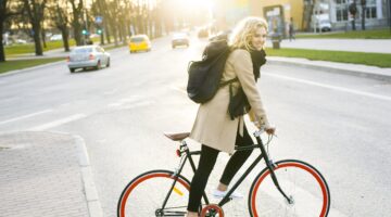 Eine lächelnde junge Frau mit Rucksack und beigem Mantel sitzt auf einem schwarzen Fahrrad mit roten