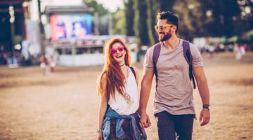 Ein junges Pärchen läuft Hand in Hand über ein Musikfestival-Gelände, beide lachen und tragen Sonnen