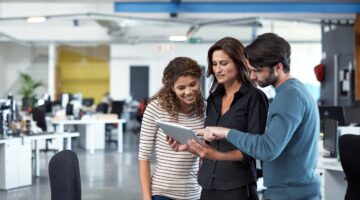 Drei junge Berufstätige stehen in einem modernen Open-Space-Büro zusammen und schauen gemeinsam auf