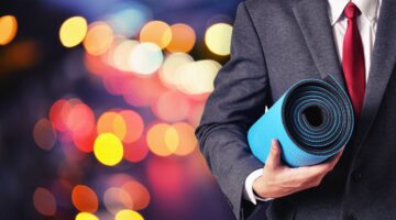 Ein Mann im grauen Anzug mit roter Krawatte hält eine zusammengerollte blaue Yogamatte unter dem Arm