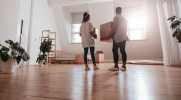 Ein junges Pärchen trägt gemeinsam einen Karton in eine helle, leere Wohnung mit Parkettboden und Zi