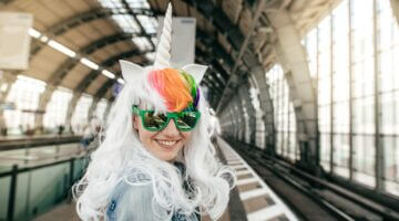 Eine lachende junge Frau in einem Einhorn-Kostüm mit weißer Perücke, buntem Regenbogenhaar, Einhorn-