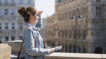 Eine junge Frau mit Jeansjacke und Umhängetasche hält eine Karte in der Hand und schaut sich suchend