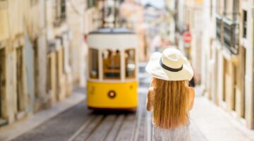 Eine junge Frau mit Strohhut und langen roten Haaren steht von hinten fotografiert in einer engen Ga